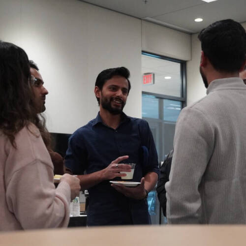 A group of people engage in a conversation in an indoor space. The person in the center, holding a drink and a plate, smiles while speaking with others gathered around.